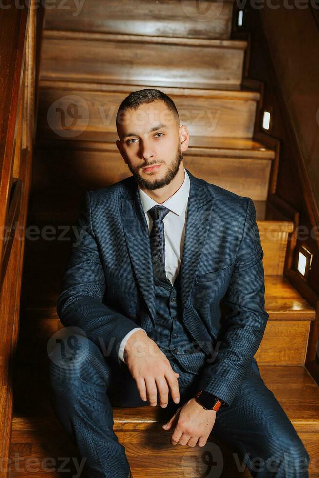 elegant groom with a beard in a beautiful blue jacket and white shirt. Businessman in classic clothes. Brunette man sits on wooden stairs. Studio portrait photo