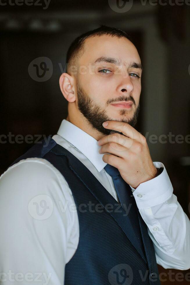 elegant groom with a beard in a beautiful blue jacket and white shirt. Businessman in classic clothes. A brunette man prepares for the wedding in the morning on his wedding day. photo