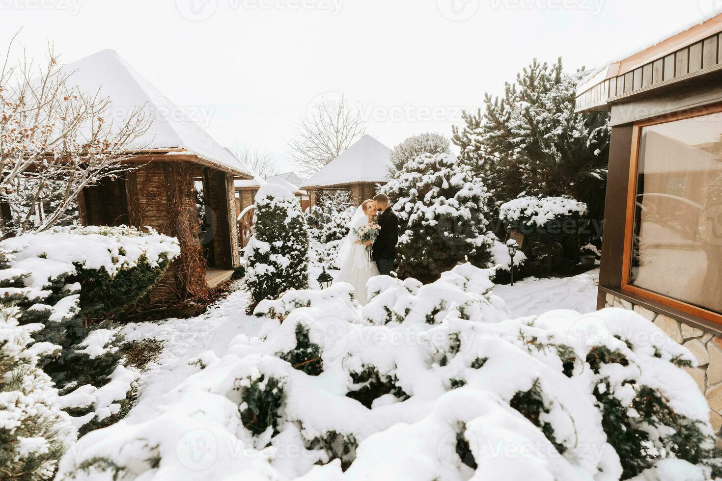 happy newlyweds between snowy trees. The groom hugs the bride in the winter park. Smiling bride with a bouquet of flowers in a wedding dress and white poncho. The groom is dressed in a black coat. photo