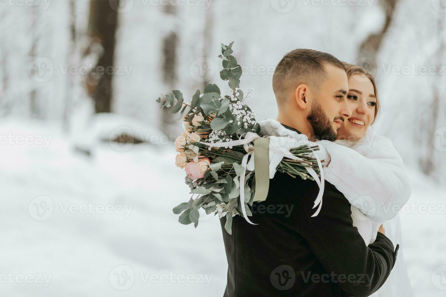 Wedding in winter. The bride and groom are standing against a snowy forest with a blurred background. The bride hugs the groom while holding a bouquet of flowers photo