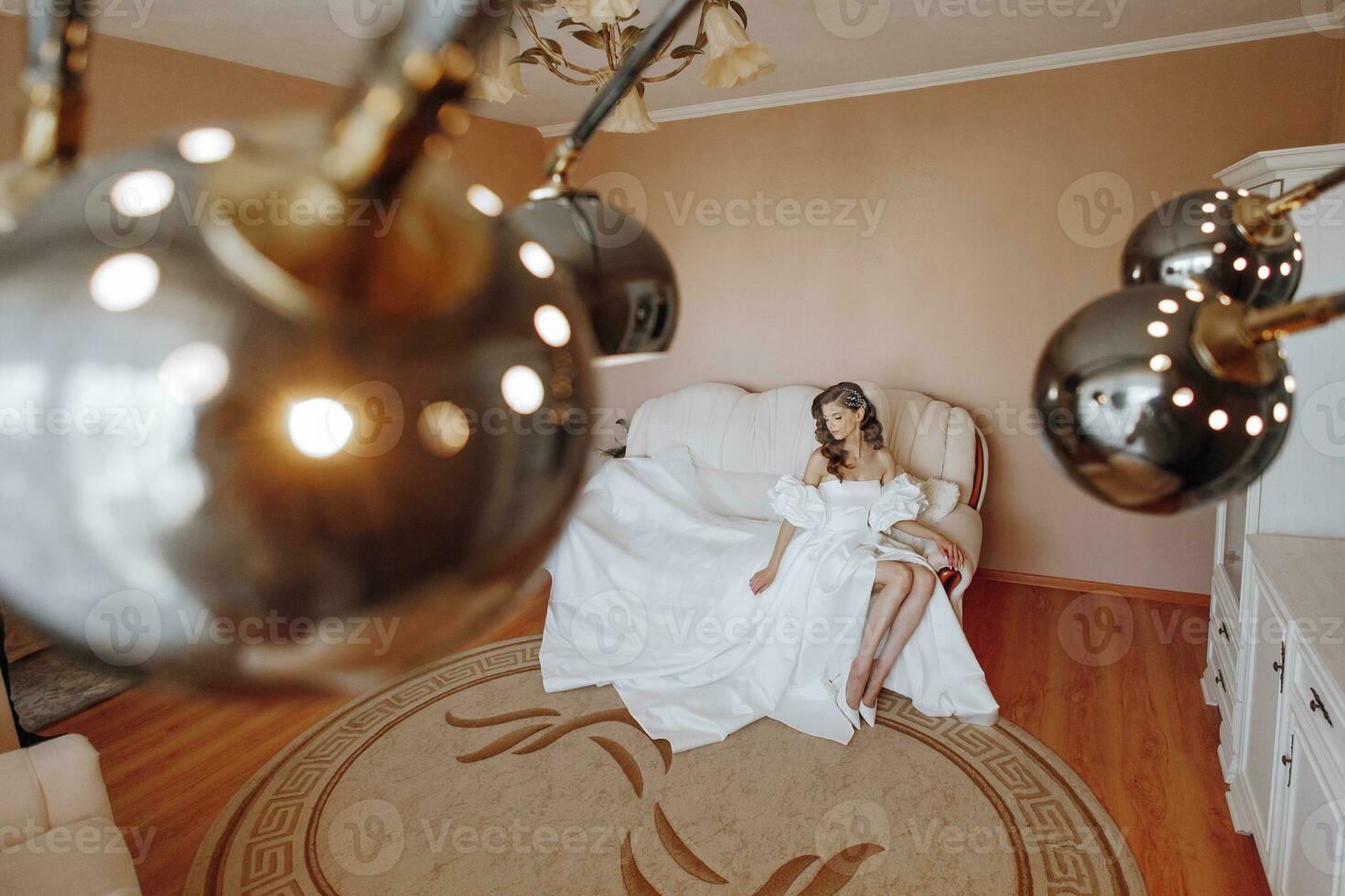 the bride in a wedding dress is sitting on the sofa. Beautiful model girl with long curly hair in a fashionable off-the-shoulder wedding dress photo