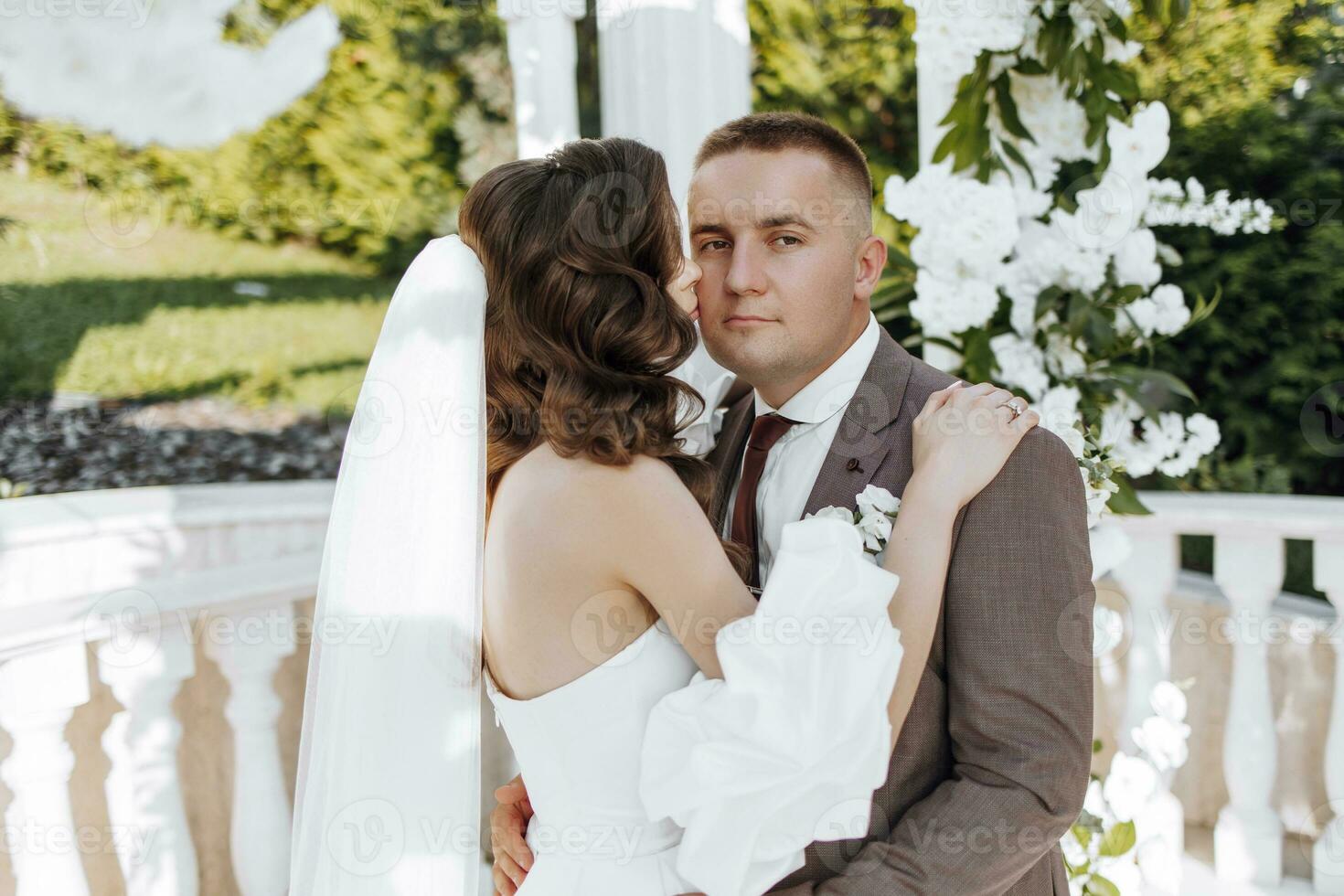 An elegant bride and groom pose together outdoors on a sunny wedding day against a background of flowers in a beautiful location. The groom gently hugs the bride and kisses her. photo