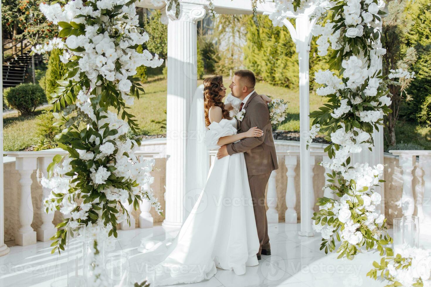 An elegant bride and groom pose together outdoors on a sunny wedding day against a background of flowers in a beautiful location. The groom gently hugs the bride and kisses her. photo