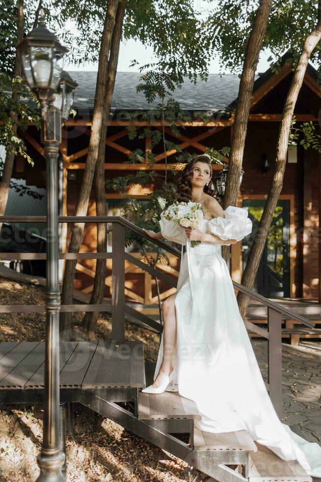 contento joven mujer con largo Rizado pelo en un blanco Boda vestido, participación un ramo de flores de flores, al aire libre. hermosa niña con flores vertical foto