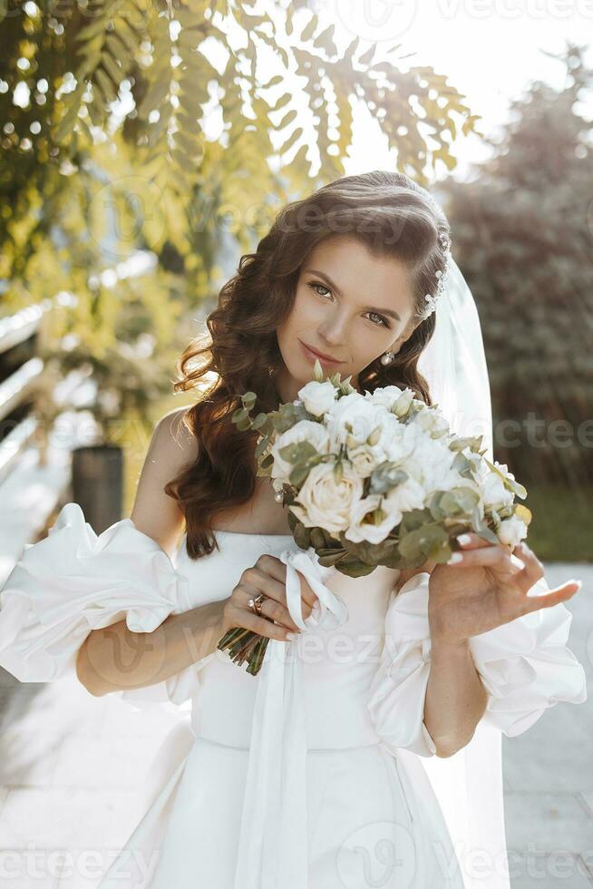 Happy young woman with long curly hair in a white wedding dress, holding a bouquet of flowers, outdoors. Beautiful girl with flowers. Vertical photo