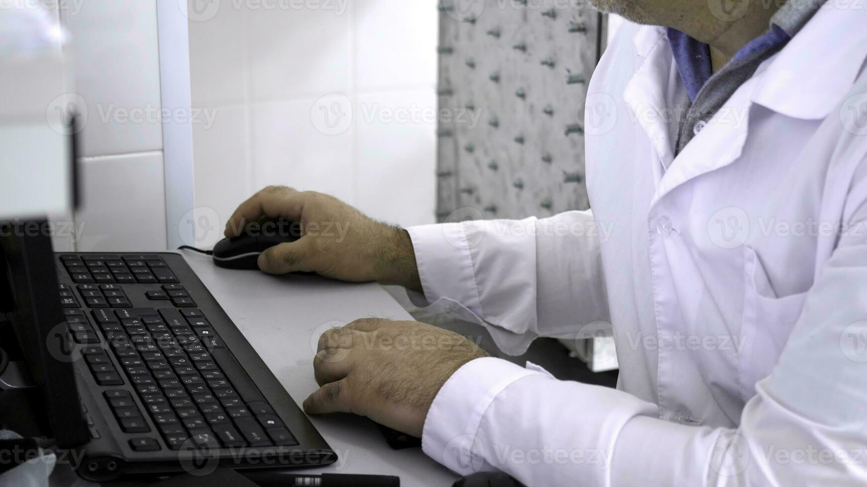 Close-up of man in white coat sitting at computer. Man in white coat sitting at computer in white room is doctor or scientist photo