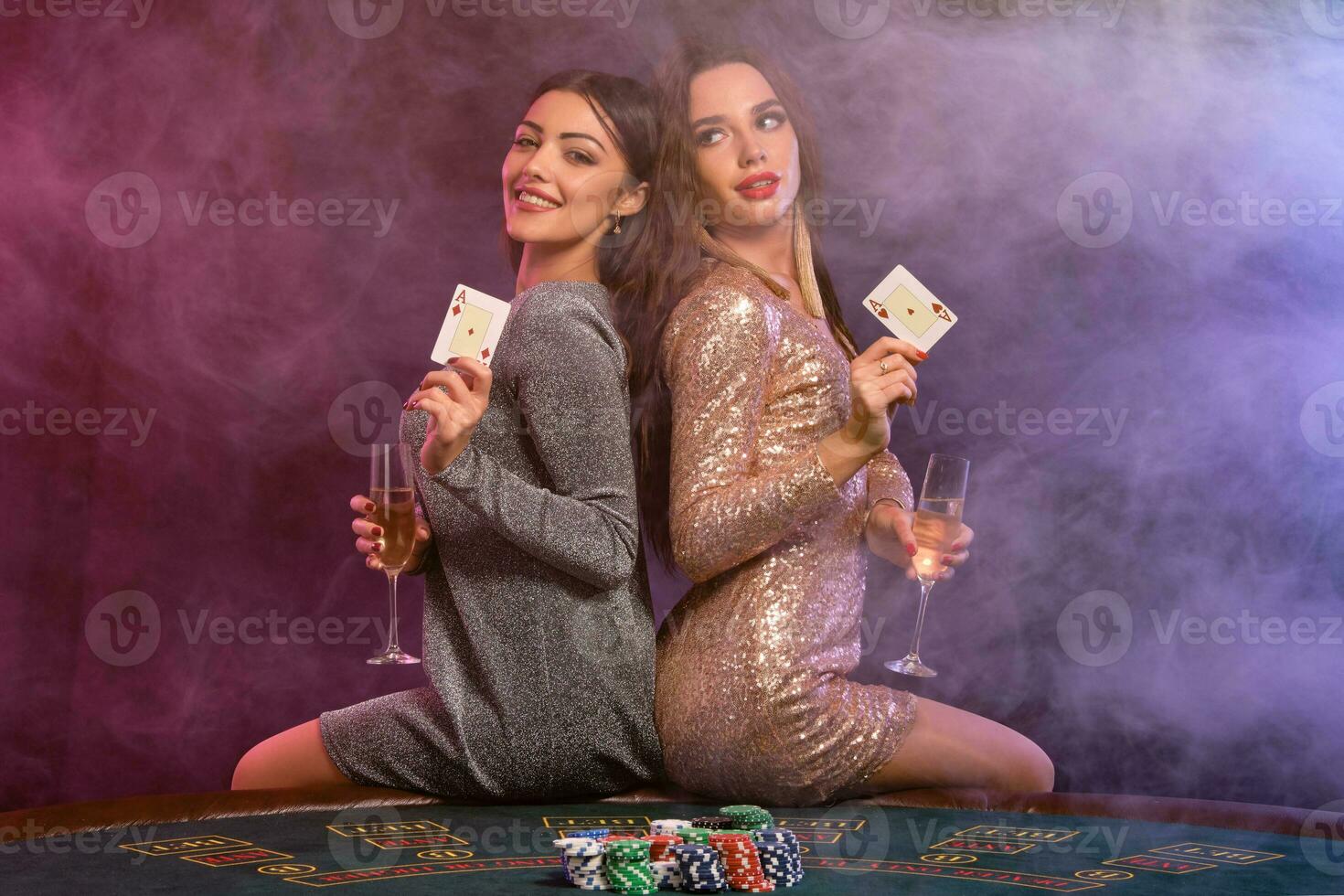 Two girls are playing poker at casino, holding glasses of champagne and cards, sitting on table with chips on it. Black, smoke background. Close-up. photo