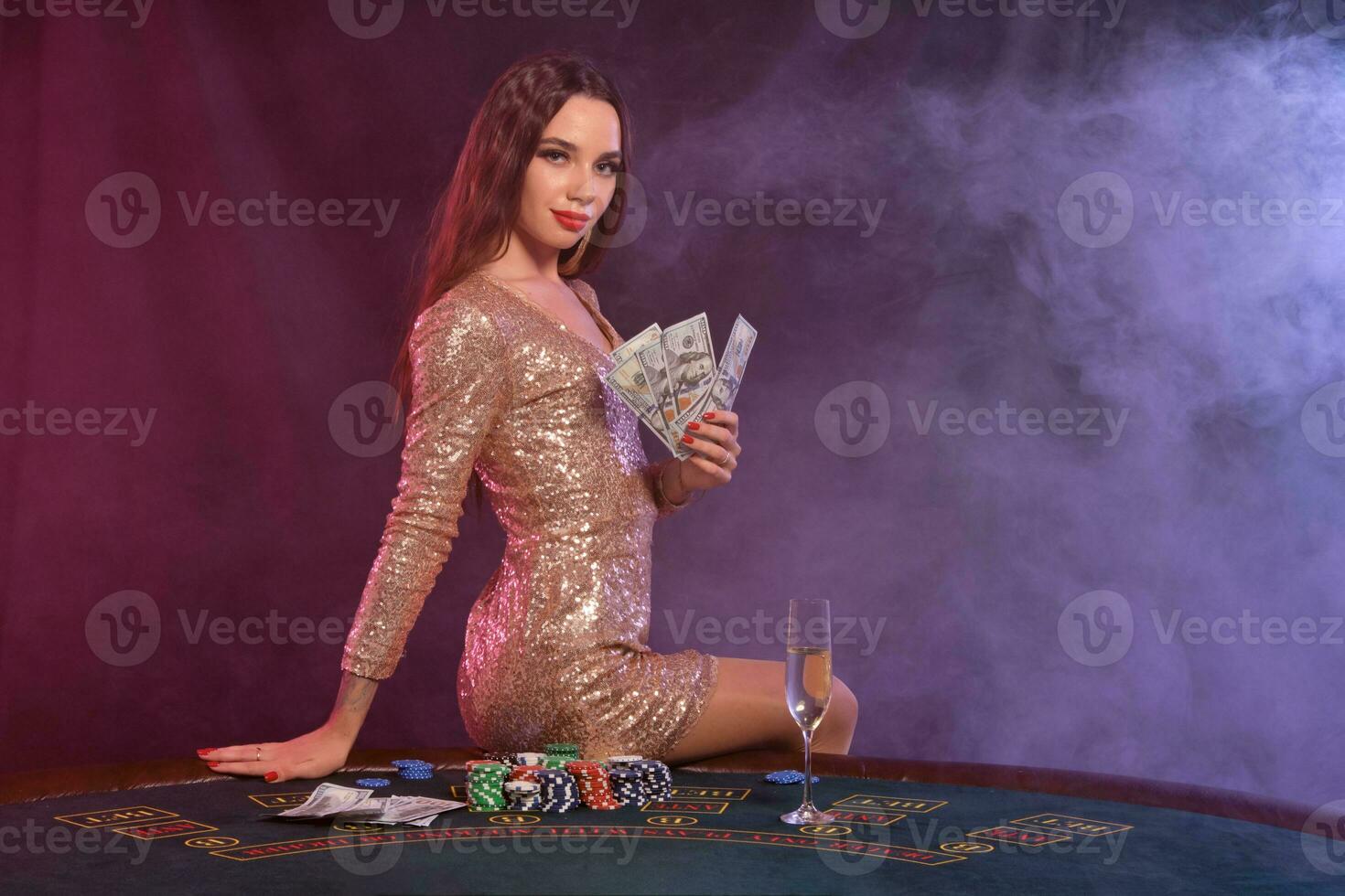 Girl playing poker at casino, holding money. Sitting on table with chips, champagne, cash on it. Black, smoke background. Close-up. photo