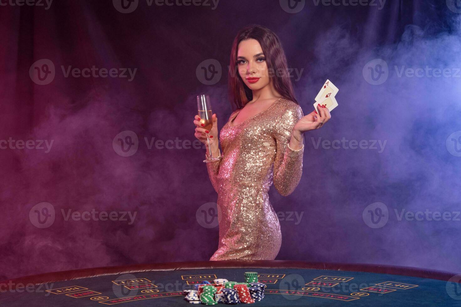 Girl playing poker at casino, holding glass of champagne, two aces. Posing at table with chips, money on it. Black, smoke background. Close-up. photo