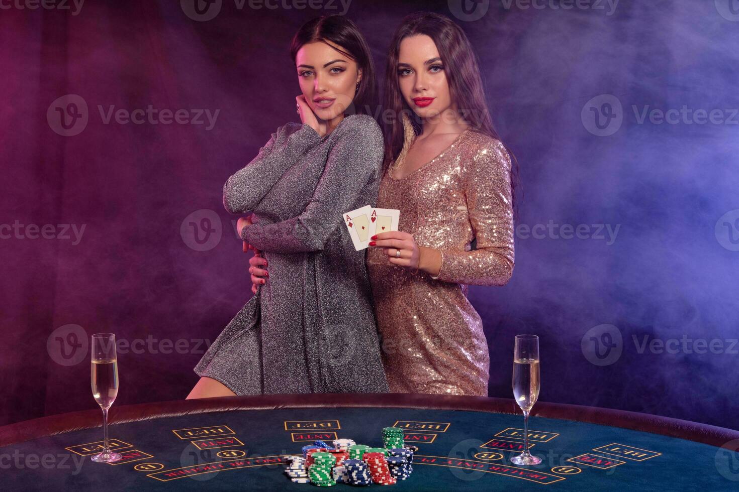 Two girls are playing poker at casino, hugging, holding cards, posing at a table with chips, champagne on it. Black, smoke background. Close-up. photo