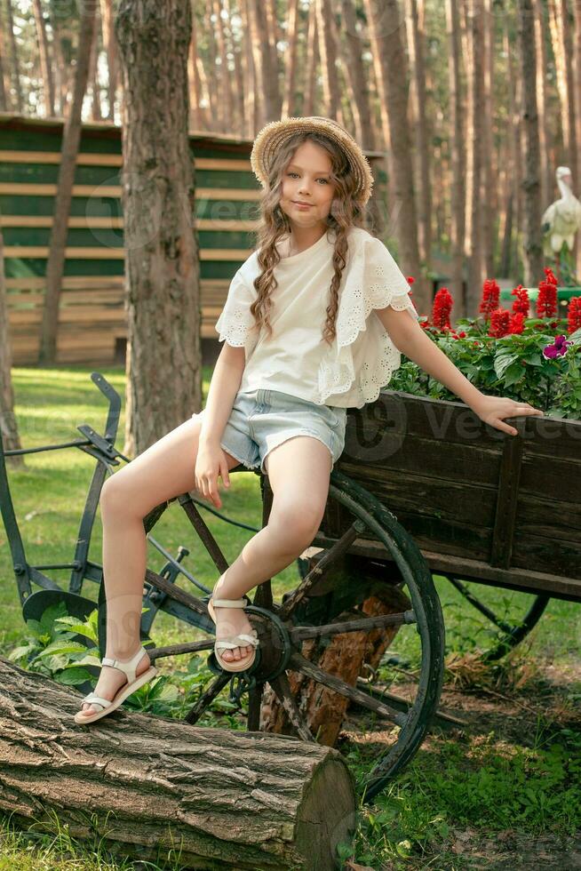 Carefree tween girl sitting on old wooden cart decorated as flowerbed at country estate in forest photo