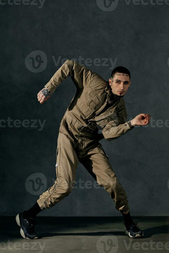 Young fellow with tattooed body and face, earrings, beard. Dressed in khaki jumpsuit, black sneakers. Dancing on gray background. Dancehall, hip-hop photo