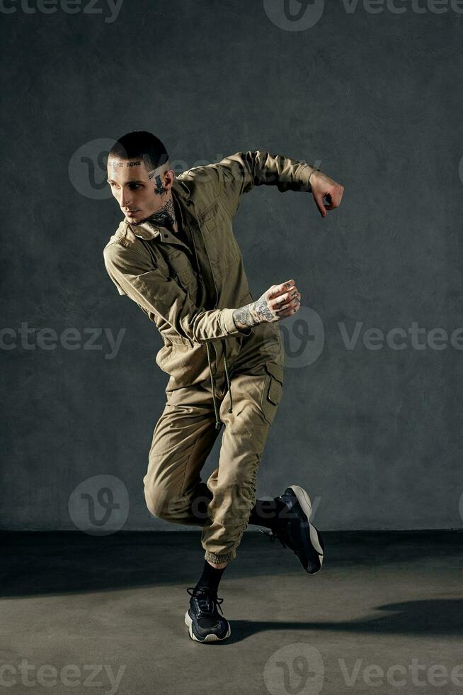Modern guy with tattooed body and face, earrings, beard. Dressed in khaki overalls and black sneakers. Dancing on gray background. Dancehall, hip-hop photo