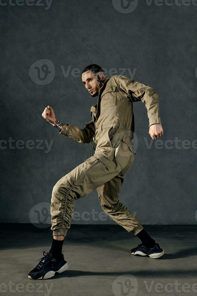 Good-looking man with tattooed body, earrings, beard. Dressed in khaki overalls and black sneakers. Dancing on gray background. Dancehall, hip-hop photo