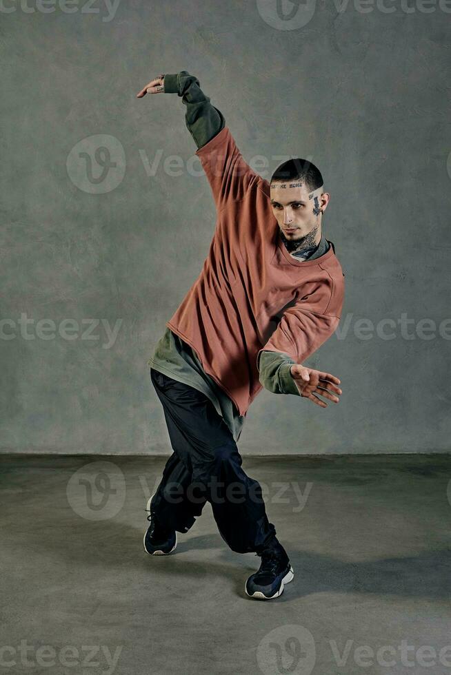 majestuoso compañero con tatuado rostro, barba. vestido en vistoso saltador, negro pantalones y zapatillas. baile, gris antecedentes. Baile (salón, hip hop foto