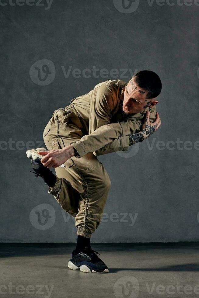 Attractive man with tattooed body and face, earrings, beard. Dressed in khaki overalls, black sneakers. Dancing on gray background. Dancehall, hip-hop photo
