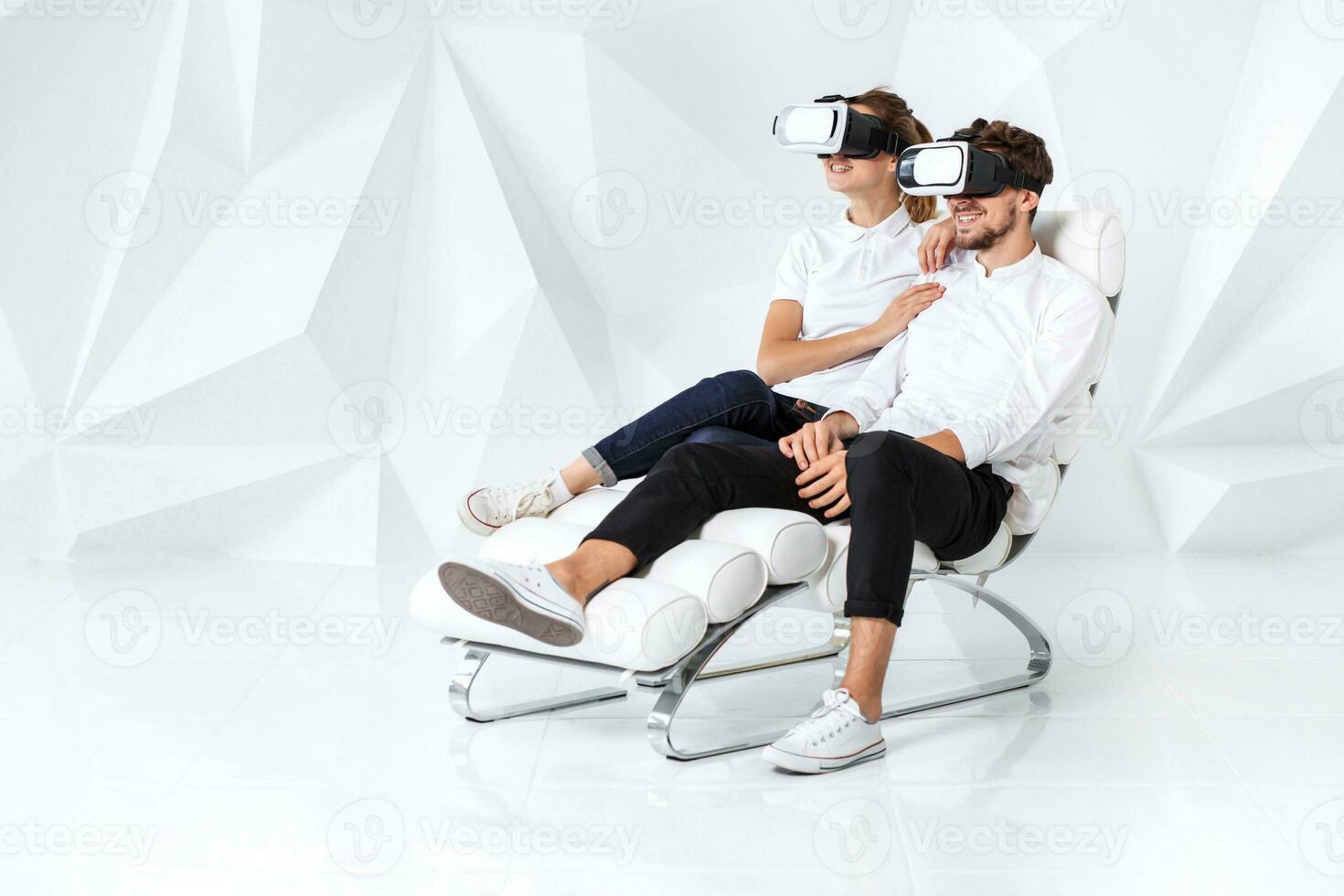 A young couple wearing VR headsets sitting on white chair in a room with white walls and floors photo