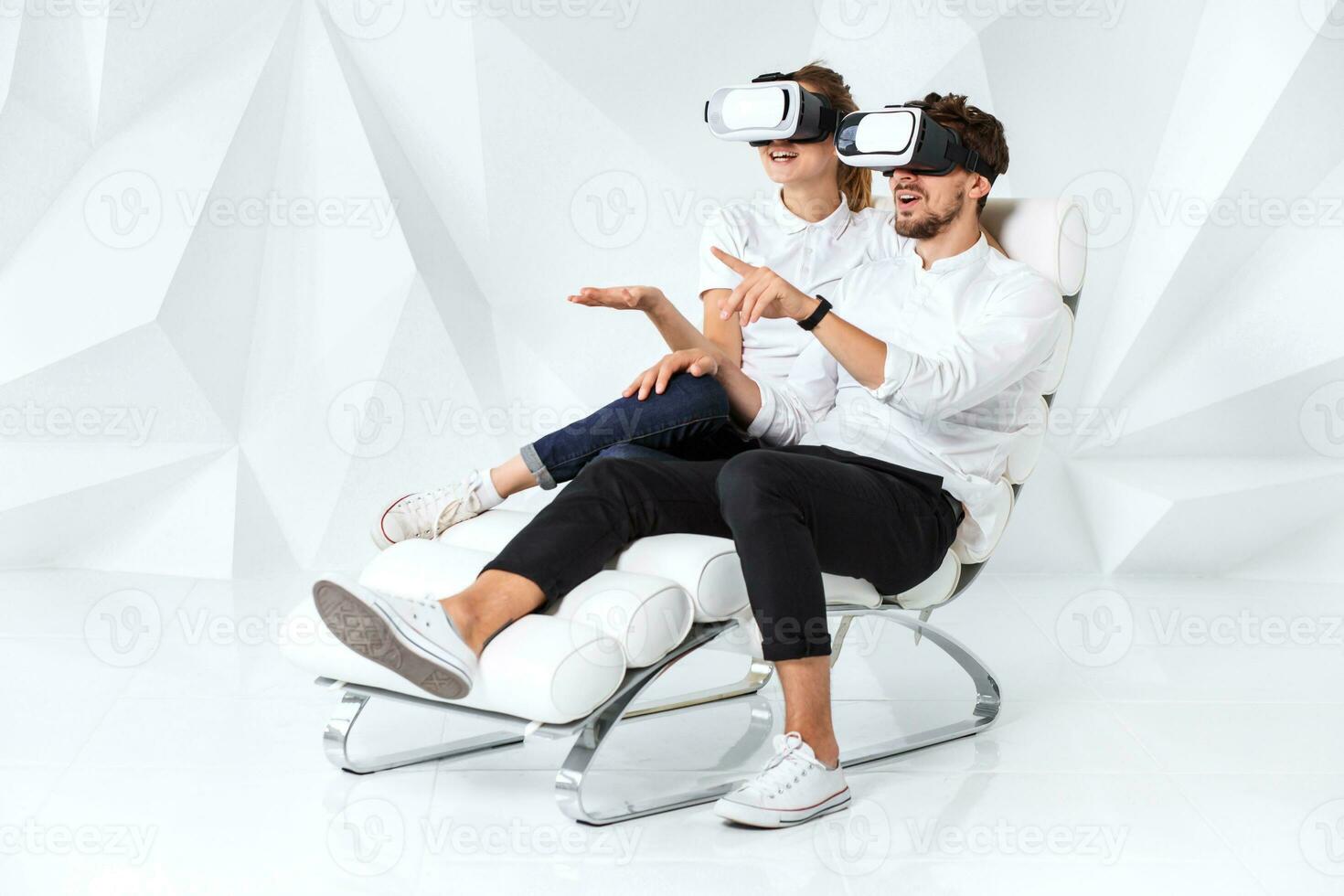 A young couple wearing VR headsets sitting on white chair in a room with white walls and floors photo
