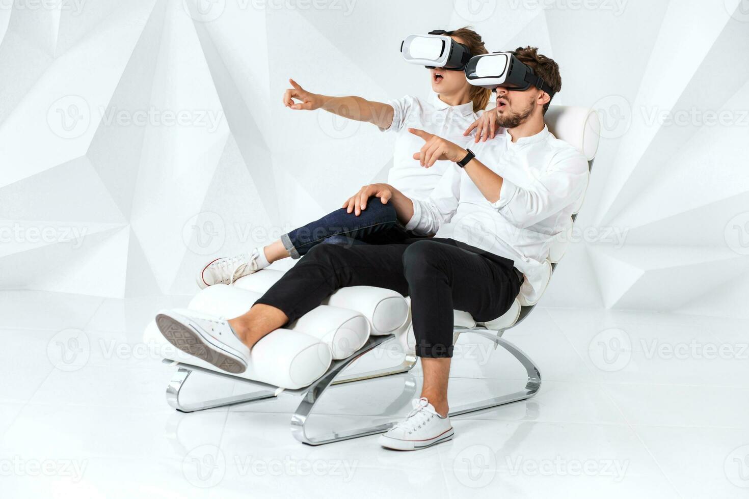 A young couple wearing VR headsets sitting on white chair in a room with white walls and floors photo