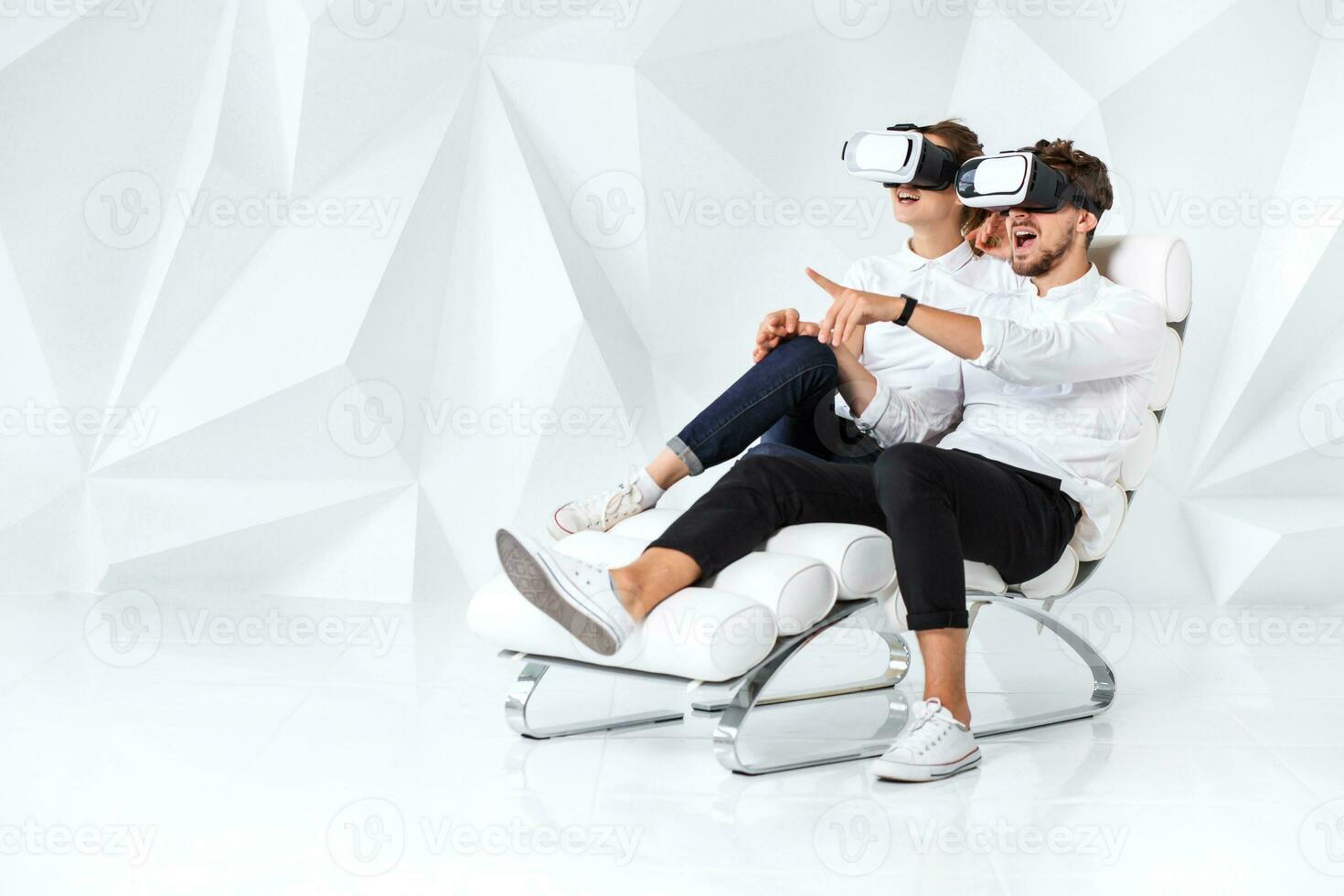 A young couple wearing VR headsets sitting on white chair in a room with white walls and floors photo