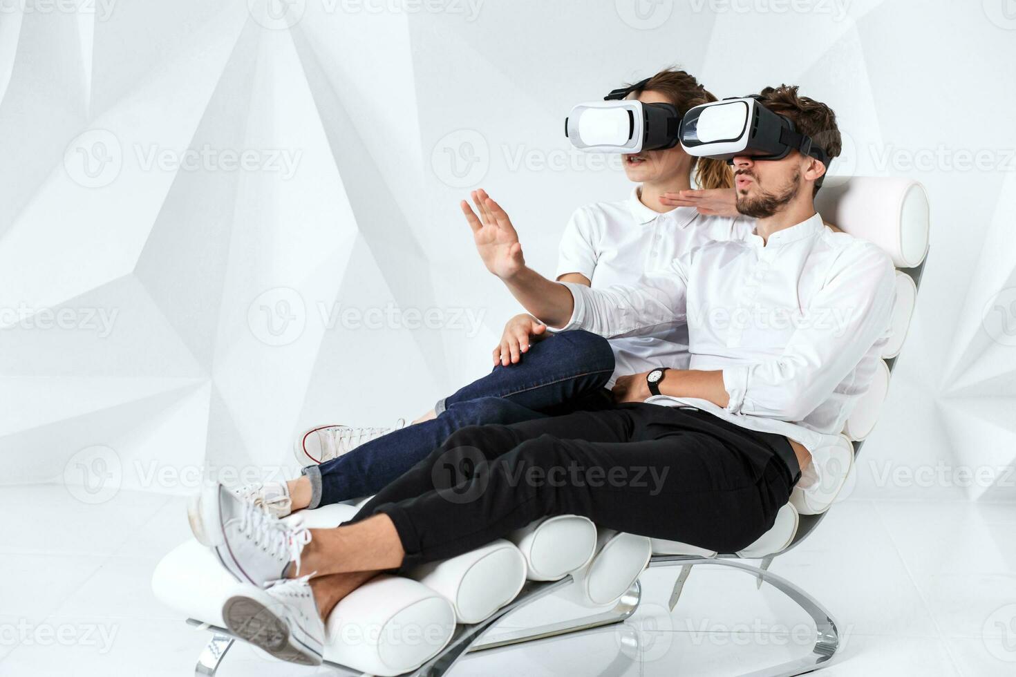 A young couple wearing VR headsets sitting on white chair in a room with white walls and floors photo