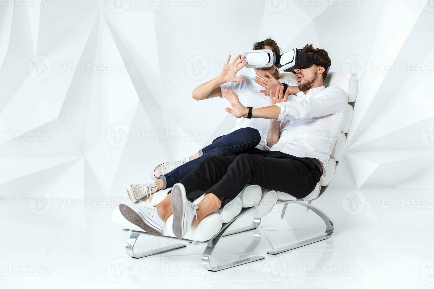 A young couple wearing VR headsets sitting on white chair in a room with white walls and floors photo