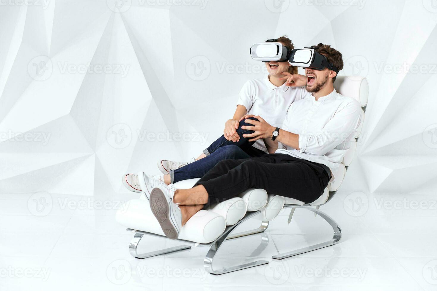 A young couple wearing VR headsets sitting on white chair in a room with white walls and floors photo