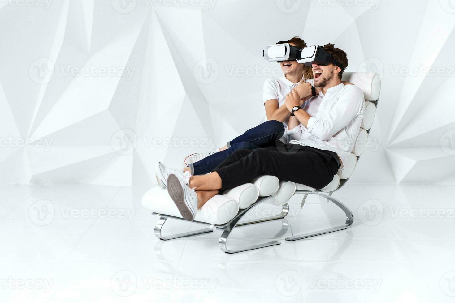 A young couple wearing VR headsets sitting on white chair in a room with white walls and floors photo