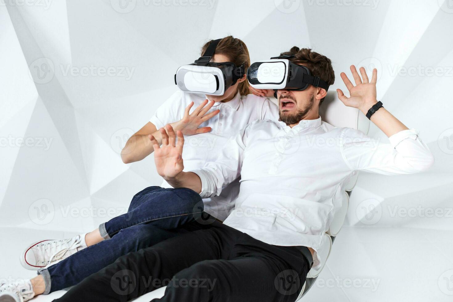 A young couple wearing VR headsets sitting on white chair in a room with white walls and floors photo