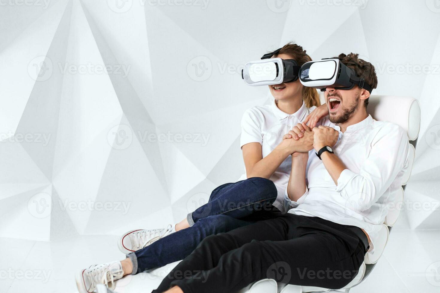 A young couple wearing VR headsets sitting on white chair in a room with white walls and floors photo