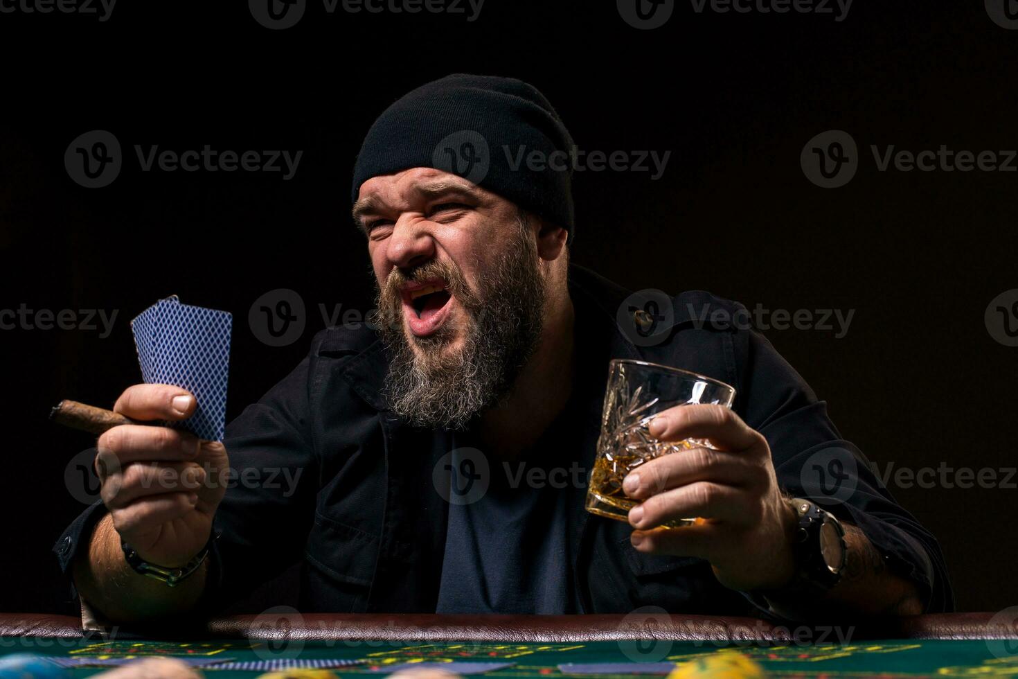Bearded man with cigar and glass sitting at poker table and screaming isolated on black photo