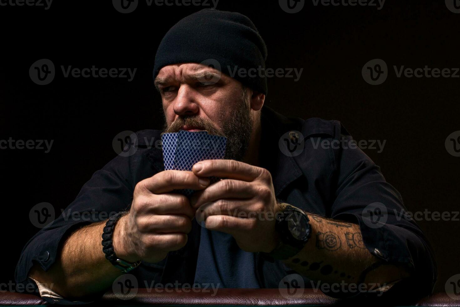 Serious bearded man sitting at poker table and holding cards isolated on black photo