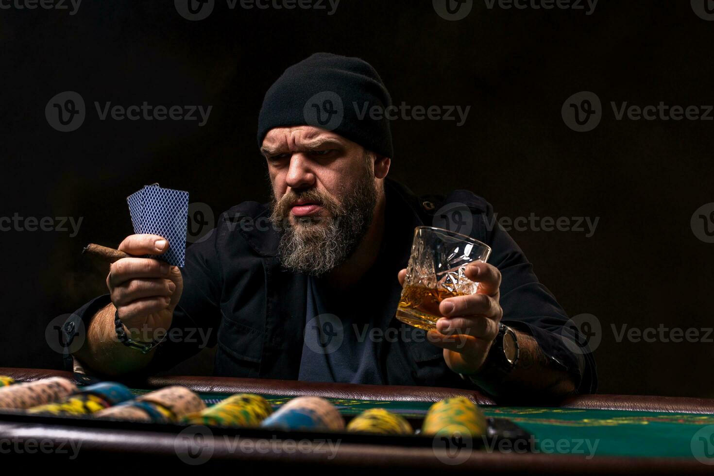 Serious bearded man with cigar and glass sitting at poker table and screaming isolated on black photo