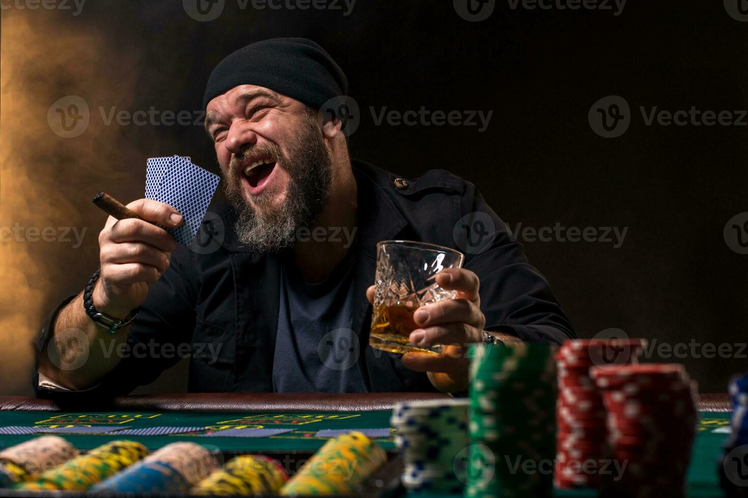 Bearded man with cigar and glass sitting at poker table and screaming isolated on black photo