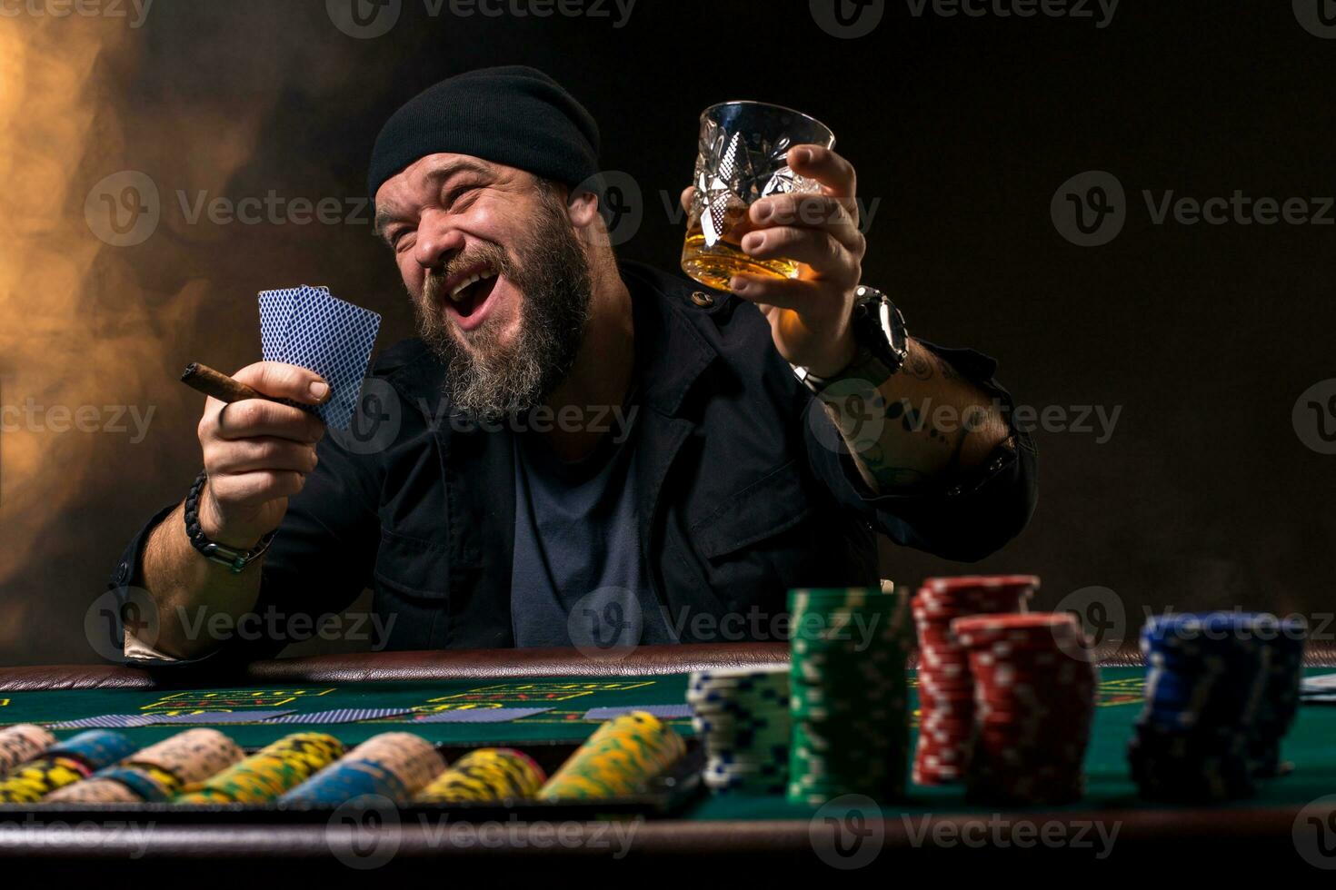 Bearded man with cigar and glass sitting at poker table and screaming isolated on black photo