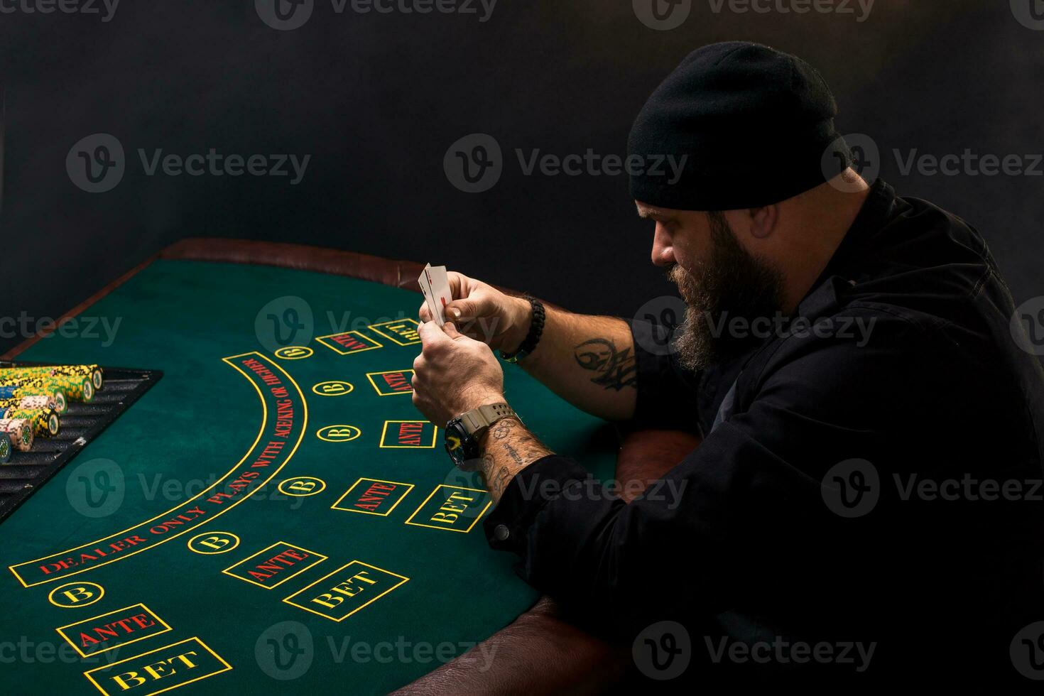 Serious bearded man sitting at poker table and holding cards isolated on black. Copy space photo