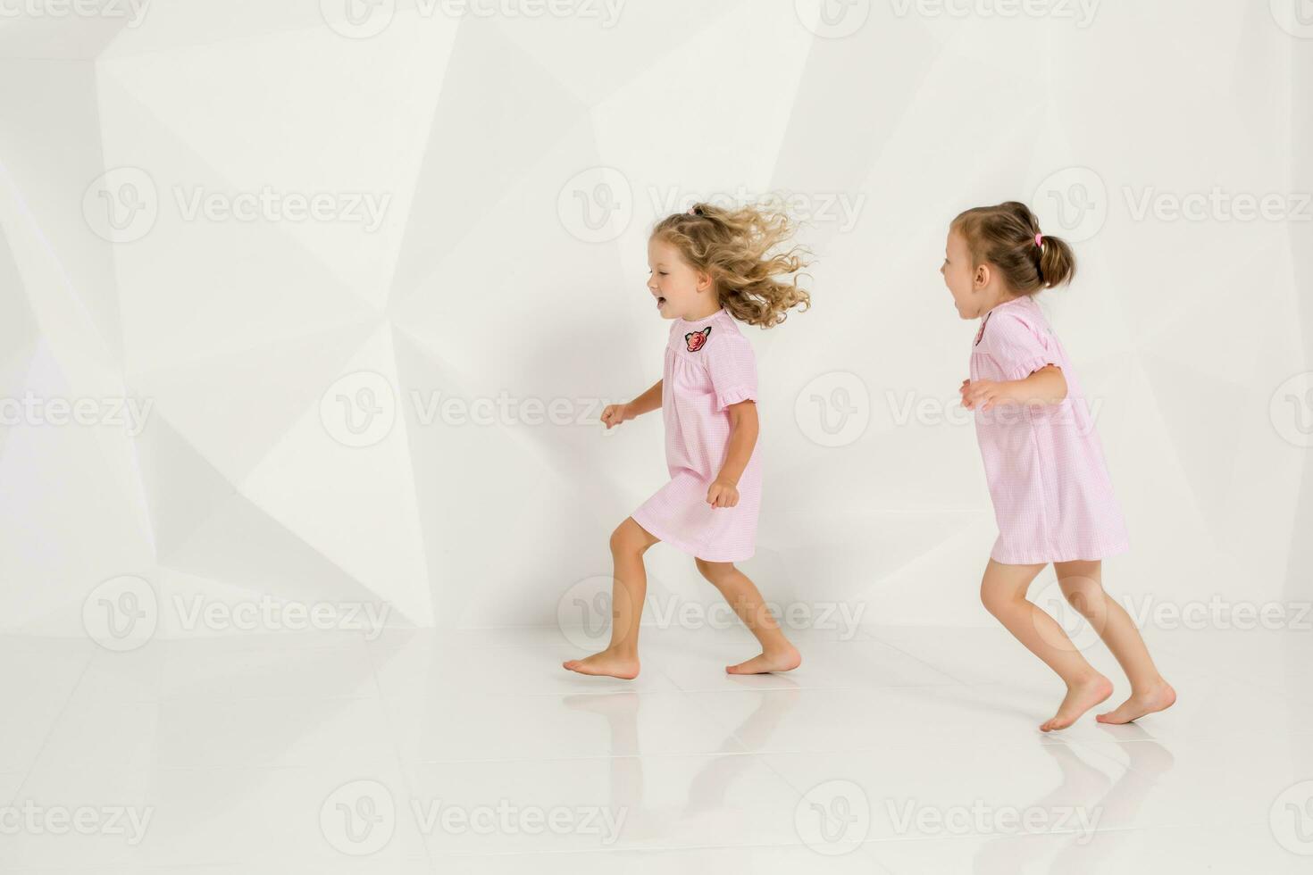 dos pequeño gracioso y riendo niña en suavemente rosado vestidos jugando en blanco estudio foto
