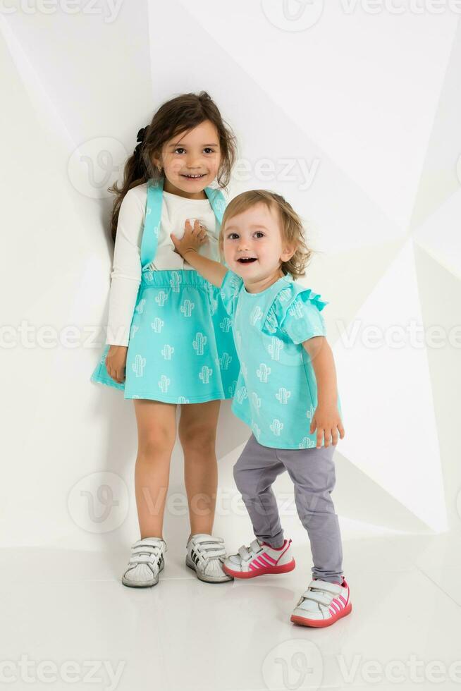 dos linda pequeño muchachas en pie en turquesa vestir en el blanco pared antecedentes en el estudio. foto