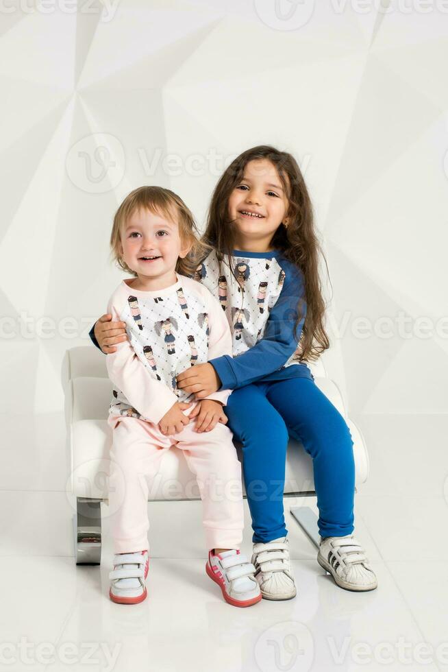 Two stylish little girls sit on one round chair on white wall background photo