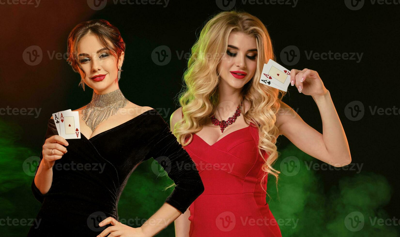 Two women in red and black dresses, jewelry. They smiling, showing playing cards, posing against smoky colorful background. Poker, casino. Close-up photo