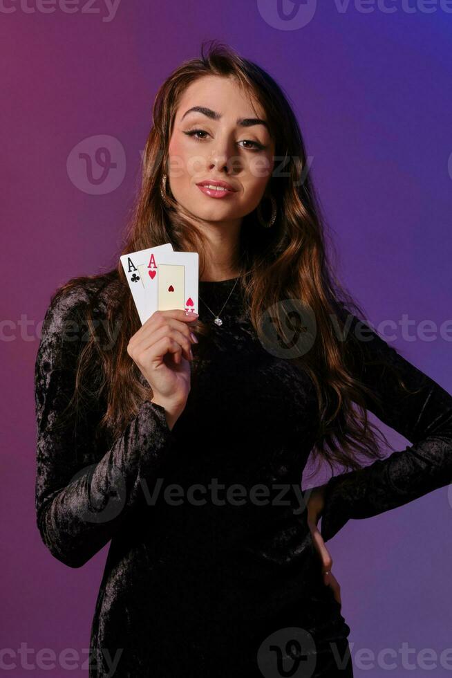 Brunette woman in black velvet dress showing two playing cards, posing against coloful background. Gambling entertainment, poker, casino. Close-up. photo