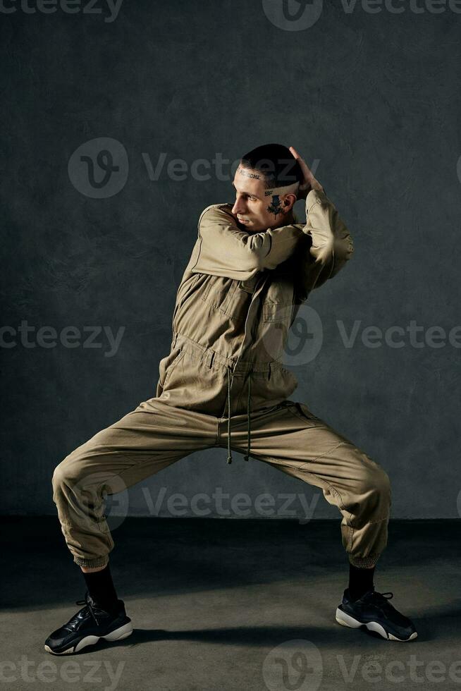 Graceful performer with tattooed body, earrings, beard. Dressed in khaki overalls and black sneakers. Dancing on gray background. Dancehall, hip-hop photo