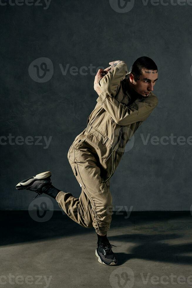 Handsome guy with tattooed body and face, earrings, beard. Dressed in khaki overalls, black sneakers. Dancing on gray background. Dancehall, hip-hop photo