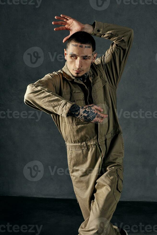 Modern male with tattooed body and face, earrings, beard. Dressed in khaki jumpsuit and black sneakers. Dancing on gray background. Dancehall, hip-hop photo