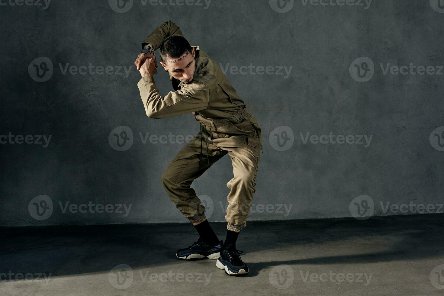 Graceful guy with tattooed body and face, earrings, beard. Dressed in khaki overalls, black sneakers. Dancing on gray background. Dancehall, hip-hop photo