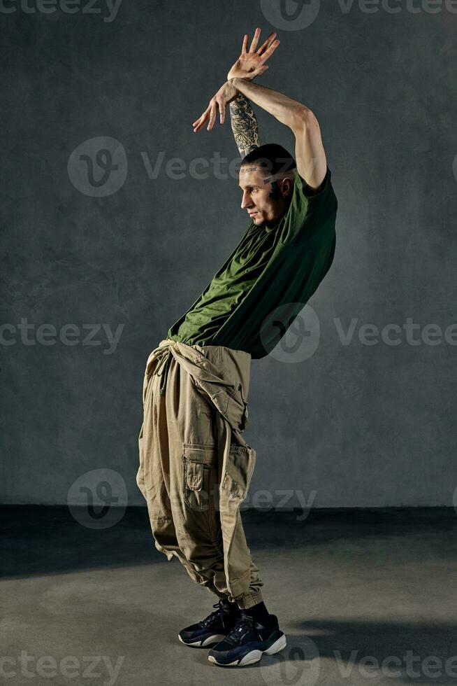 Performer with tattooed body, earrings, beard. Dressed in khaki t-shirt and overalls, black sneakers. Dancing on gray background. Dancehall, hip-hop photo