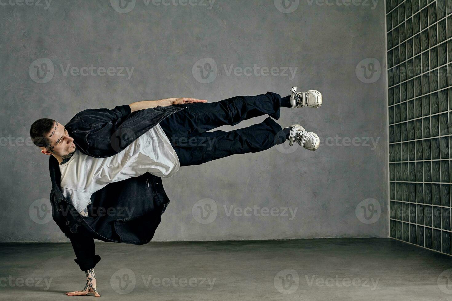 Guy with tattooed body, beard. Dressed in white t-shirt and sneakers, black shirt, pants. Showing tricks on gray background. Dancehall, hip-hop photo