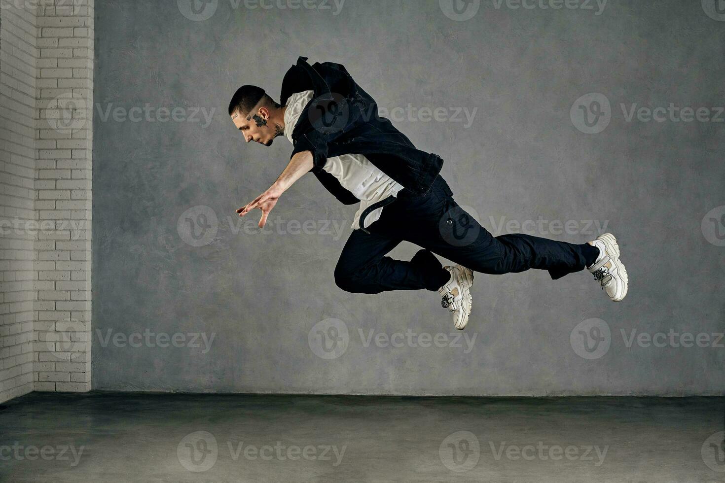 Guy with tattooed body, beard. Dressed in white t-shirt and sneakers, black shirt, pants. Performing tricks on gray background. Dancehall, hip-hop photo