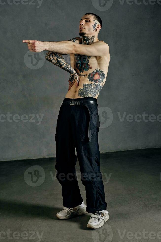 Flexible tattooed guy with naked torso, beard. Dressed in black pants and white sneakers. Dancing against gray background. Dancehall, hip-hop photo