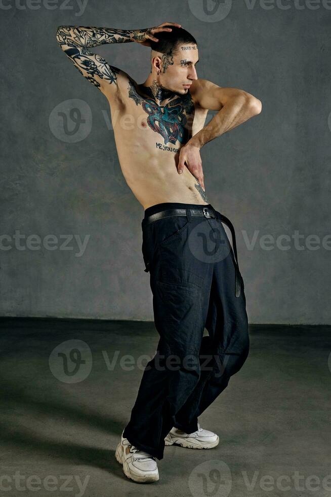 Flexible tattooed performer with naked torso, beard. Dressed in black pants and white sneakers. Dancing against gray background. Dancehall, hip-hop photo